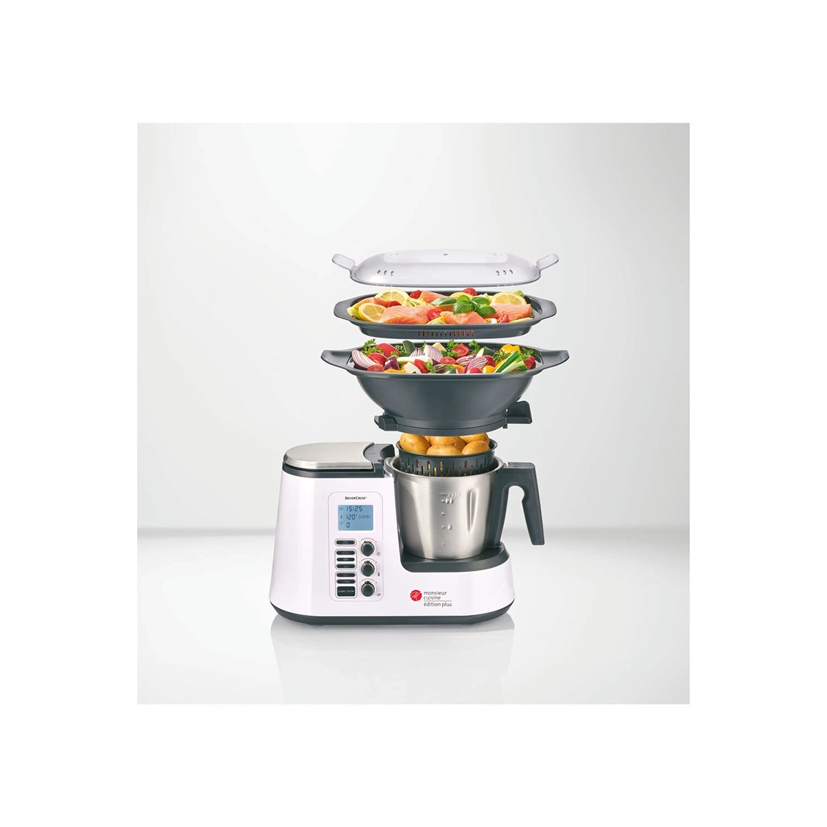 Gezeigt wird eine SILVERCREST Monsieur Cuisine edition plus SKMK 1100 A1 Küchenmaschine in Weiß mit digitalem Display, mit herausnehmbaren, gestapelten und mit verschiedenen Gemüsesorten gefüllten Dampfschalen, die über dem Hauptgerät auf einem unifarbenen Hintergrund angeordnet sind.