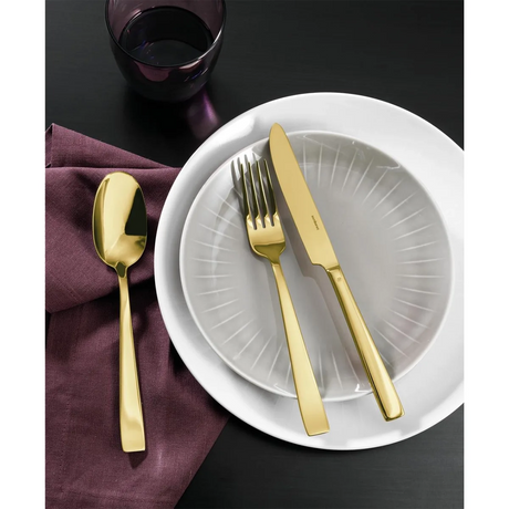Das Sambonet Flat Besteck 24tlg. - gold von Sambonet, aus Edelstahl 18/10 mit PVD-Gold-Finish, ruht auf einer violetten Serviette neben einem weißen Teller mit durchscheinendem grauen Innenteller und goldenem Besteck. Darüber steht ein dunkles Glas auf dem schwarzen Tisch.